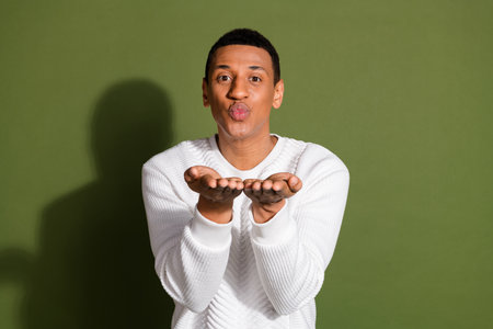 Young man blowing a playful kiss in front of khaki background wearing a white sweater, expressing...