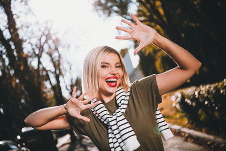 Photo of pretty young blonde woman excited have fun raise hands wear khaki clothes tied shoulders sweater walk outside urban city parkの写真素材