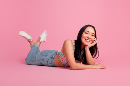Smiling young woman lying on pink background wearing casual stylish summer outfit, exuding joy and confidenceの写真素材