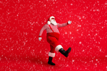Full length profile photo of grandfather grey beard step raise hand wear santa claus x-mas costume suspenders sunglass striped shirt cap boots isolated red color backgroundの写真素材