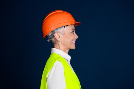 Middle-aged woman in hardhat and safety vest confidently smiles, showcasing professionalism in the...