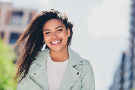 Charming young woman with a delightful smile enjoying a sunny day outdoors in the city, wearing a...