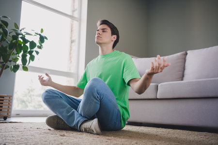 Young man meditating in living room near sofa in casual green t-shirt and jeans enjoying peaceful indoors moment with nature and daylightの写真素材