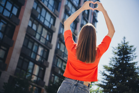 Young woman in red shirt forms heart shape with hands in sunny urban setting of city streetの写真素材