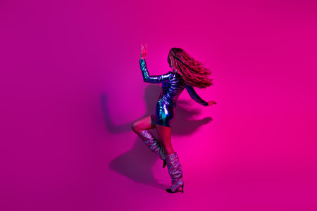 Stylish woman dancing in a shiny blue dress and boots against a bright pink background, celebrating at a nightclub party.の写真素材