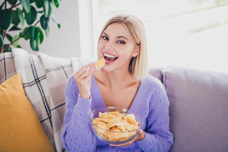Charming young woman with blond hair enjoying a snack indoors, relaxing in cozy home settingの写真素材