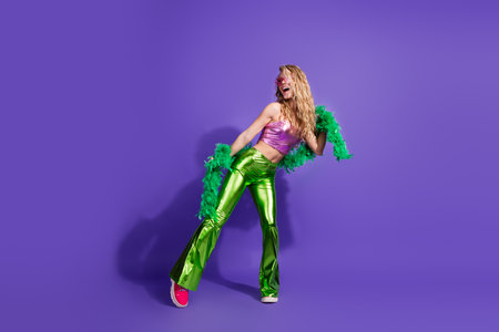 Stylish young woman in glowing top and green pants enjoying disco vibes against purple background