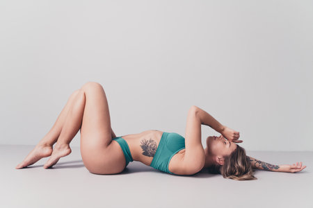 Serene young woman with natural beauty in stylish sportswear lying on a studio floor with tattoo visible, relaxing under soft, elegant daylightの写真素材