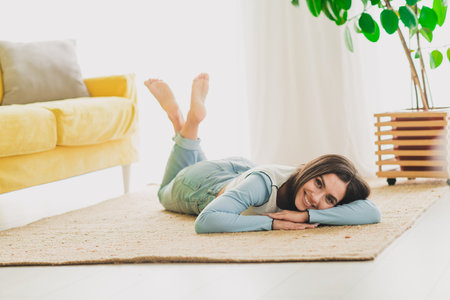 Smiling young woman lying on a cozy rug in a bright and modern living room, enjoying a moment of relaxation and comfort in daylight.の写真素材