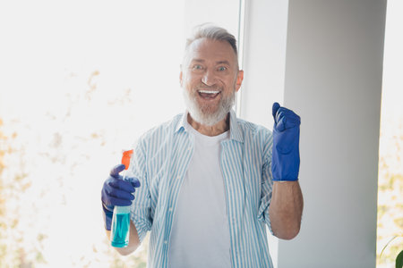 Happy senior man wearing gloves and holding a spray bottle in a brightly lit home setting, embodying positive lifestyleの写真素材