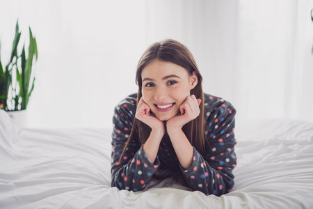 Happy young woman lying on bed in pajamas, indoors, enjoying a relaxing morning with daylight streaming into her cozy bedroomの写真素材