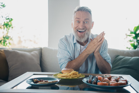 Cheerful elderly man enjoying a weekend meal at home, featuring a variety of food dishes and a comfortable living room settingの写真素材
