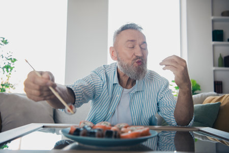 Elderly man savoring sushi at home in a casual setting, expressing joy and appreciation for the meal during a relaxed moment indoors.の写真素材