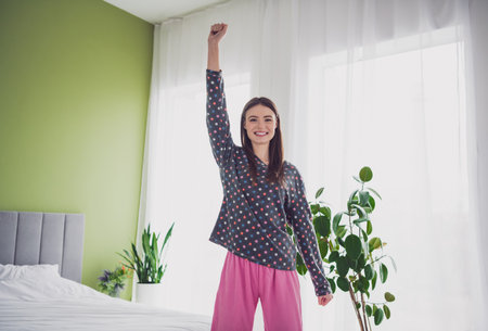 Young woman in colorful sleepwear happily raising hand in a bright bedroom with plants and natural lightingの写真素材