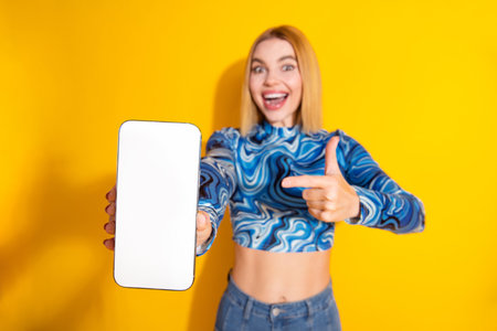 Young woman showing smartphone on vibrant yellow background promoting digital contentの写真素材