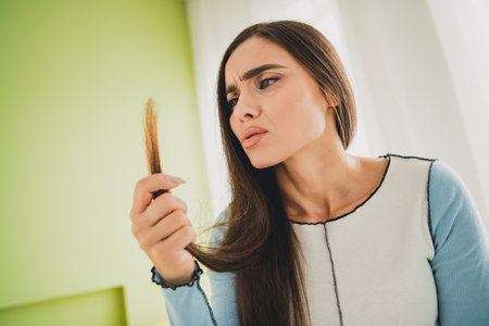 Young woman examining hair, expressing concern about its condition, observing split ends in a bright living room interiorの写真素材