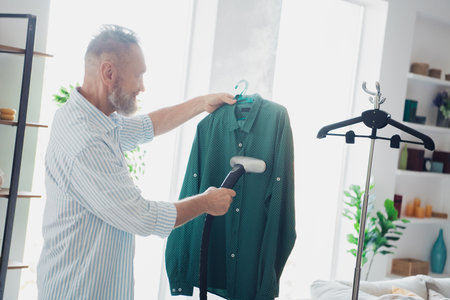 Mature man steaming a green shirt at home, combining household chores with comfort and leisure in a brightly litの写真素材