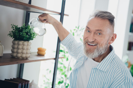 Smiling Elderly Man Watering a Plant Indoors in a Cozy Home Environment During Daylightの写真素材