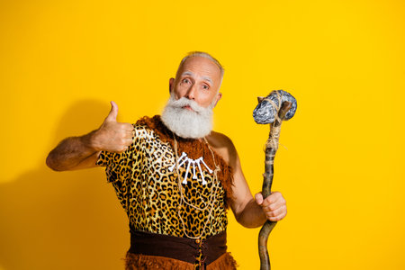 Photo of ancient aged caveman hold ax show thumb up wear leopard skin clothes isolated on yellow color backgroundの写真素材