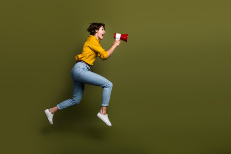 Energetic young woman holding a megaphone while leaping against an olive green background suggesting energetic promotionの写真素材