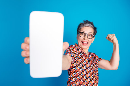 Excited mature woman showcasing smartphone with cheerful expression against vibrant blue background