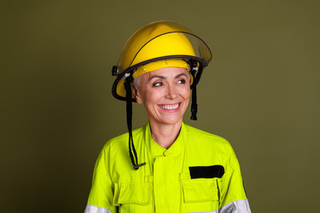 Mature female professional smiles confidently in bright, protective workwear against a khaki...