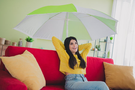 Casual young woman relaxing indoors on a colorful couch, under a large umbrella in a bright and stylish home settingの写真素材