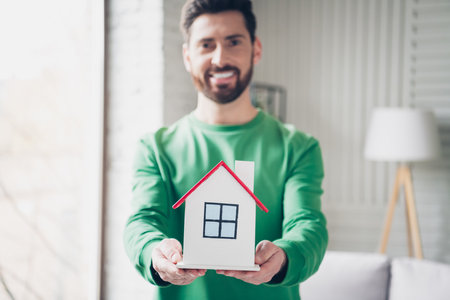 Man holding a miniature house model in a living room, symbolizing real estate, homeownership, or housing concepts.の写真素材