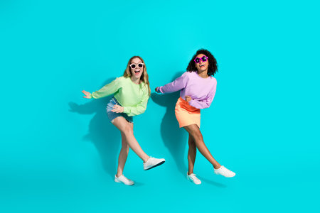 Two Trendy Young Women Posing Cheerfully Against a Bright Turquoise Backdrop, Dressed in Casual and Stylish Outfitsの写真素材
