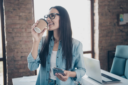 Photo of attractive young woman drink coffee device dressed denim clothes executive business leader work comfort beautiful office interiorの写真素材