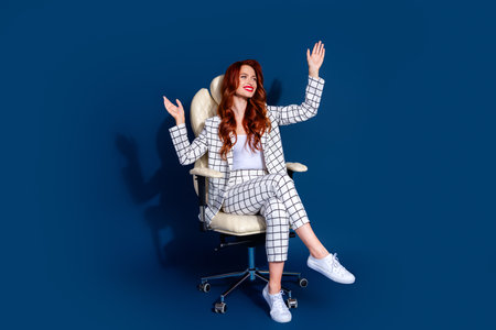 A stylish young woman with vibrant red hair sits joyfully in a chair against a dark blue background, wearing a trendy checkered suitの写真素材