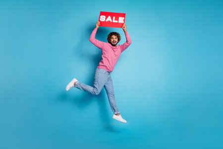 Happy young man jumping holding a sale sign, stylish casual outfit, vibrant blue background,...