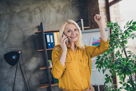 Photo of lucky positive lady broker wear shirt rising fist talking modern device indoors workplace workshopの写真素材