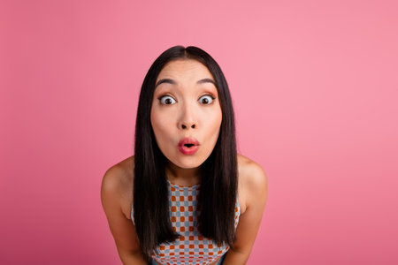 Young Asian woman in trendy outfit with surprised expression on pink background, symbolizing...