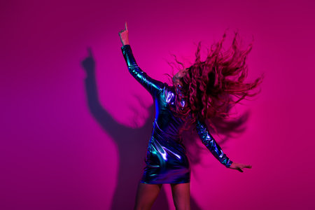 Glamorous woman in a shiny blue dress dances joyfully under vibrant pink lights, her hair flowing...