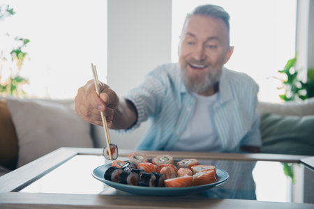 Elderly man enjoys sushi in bright daylight at home, embodying leisure and comfort, wearing casual striped shirtの写真素材