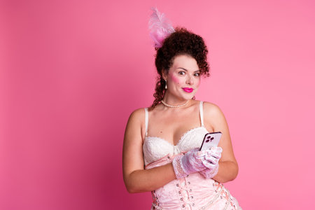 Modern twist on vintage elegance: a young woman in a classic baroque gown holding a smartphone against a vibrant pink backdropの写真素材