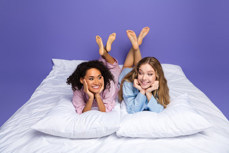 Two cheerful young women enjoying leisure time in matching pajamas on a white bed with a purple backgroundの写真素材