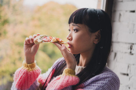 Young woman enjoying a slice of pizza with thoughtful expression beside a window on an autumn dayの写真素材