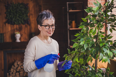 Charming elderly woman with short grey hair in a casual pullover, tending indoor plants in daylight,...