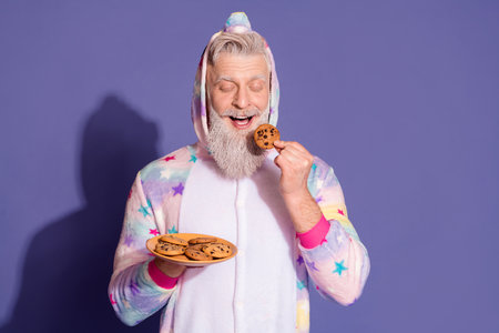 Trendy mature man in unicorn pajama enjoying cookies against a captivating purple backgroundの写真素材