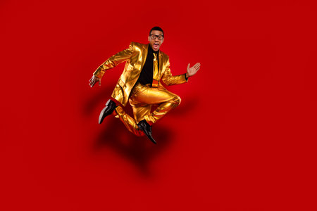 Energetic young man in a glossy gold suit jumps joyfully against a vivid red backdrop, embodying fun and style.の写真素材