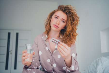 Beautiful young woman with red hair in pajamas holding medicine and water in a cozy bedroom during morning lightの写真素材
