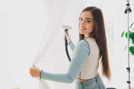 Young woman in casual attire steaming curtains at home in a bright interior, bringing comfort and charm to her living space.の写真素材