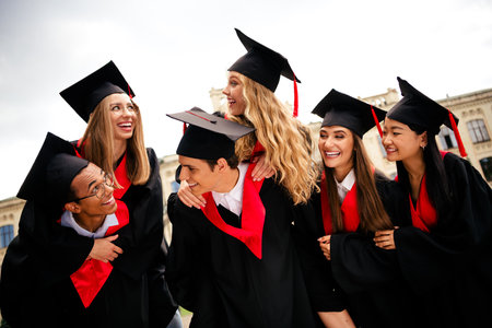 Young graduates celebrating in caps and gowns on a vibrant summer day outdoors in an urban setting, embodying joy and achievementの写真素材
