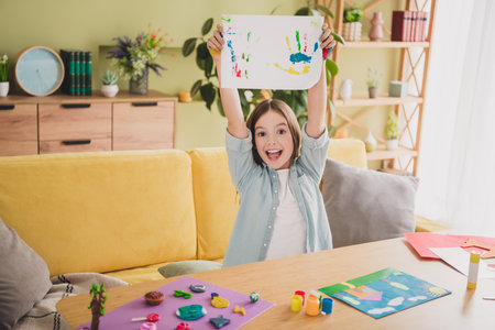Cheerful girl holding colorful handmade artwork with excitement in cozy living room