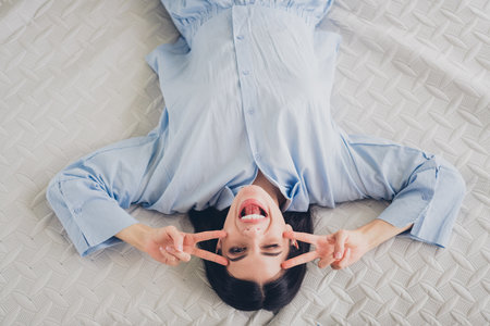 Young woman in blue pajamas joyfully waking up in bed with peace signsの写真素材