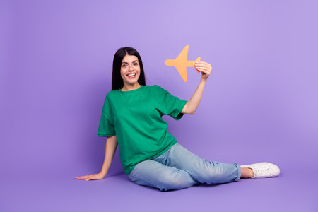 Joyful young woman in casual green t-shirt holding paper airplane with excitement against purple backdropの写真素材