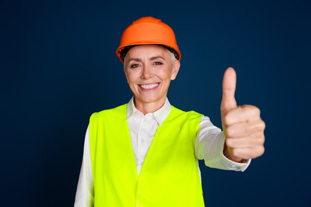 Confident mature female engineer gives a thumbs up. She stands against a dark blue background, wearing a bright safety vest and helmetの写真素材