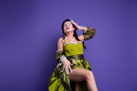 Elegant young woman in green baroque dress posing on vibrant purple background showcasing a theme of...